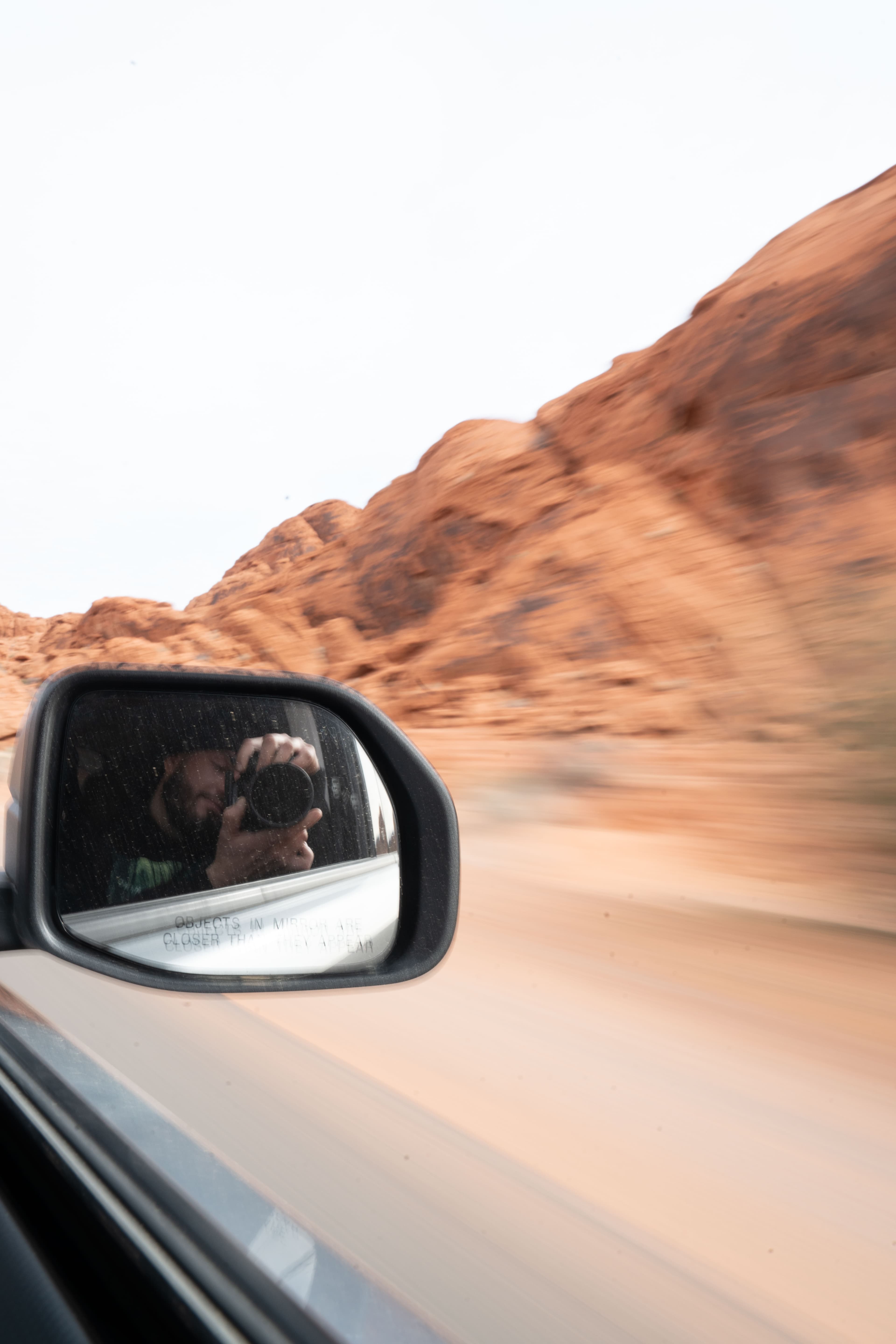 A photographer reflected in a car mirror beside red desert rock.