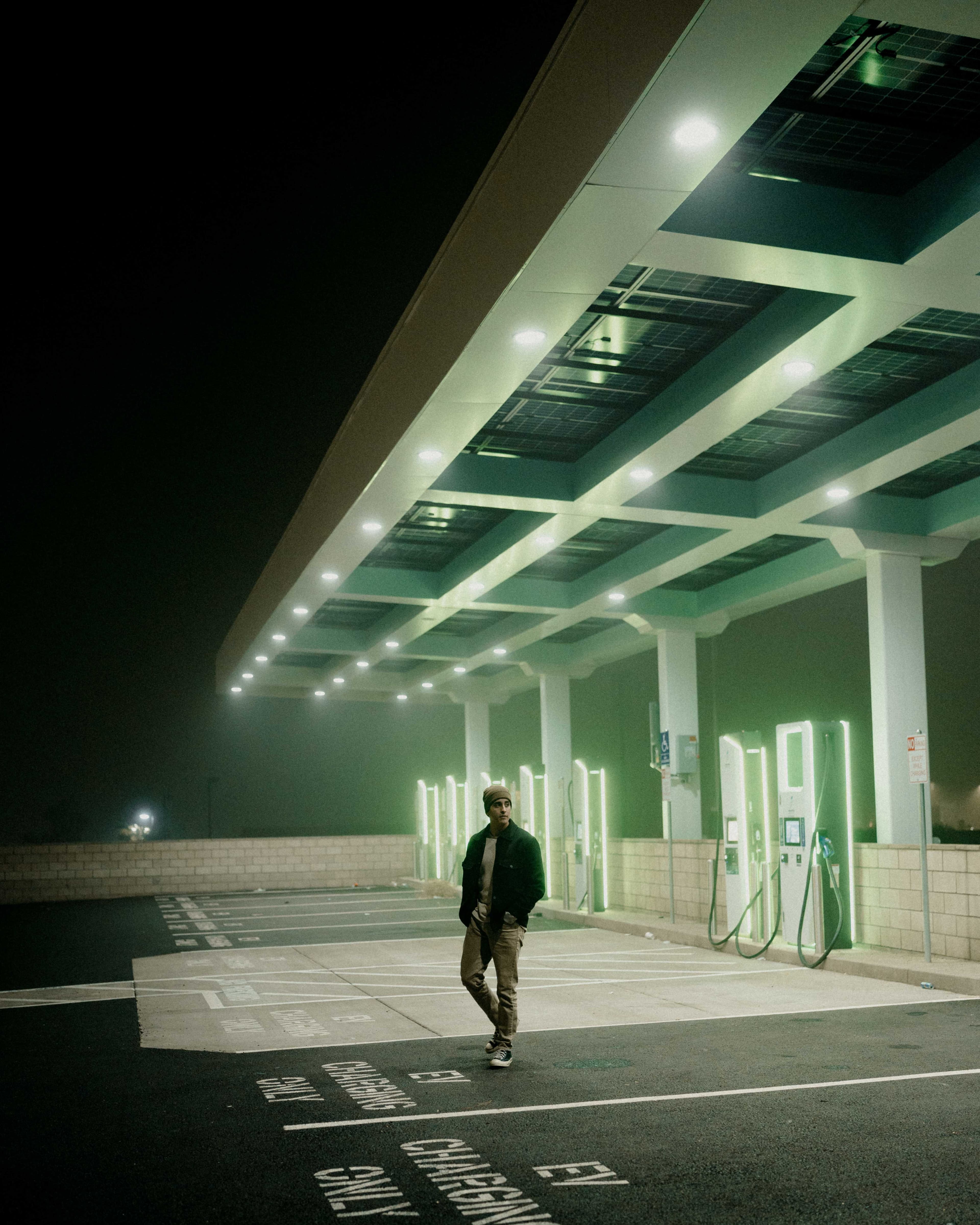 A man walking beneath green-lit charging bays at night.