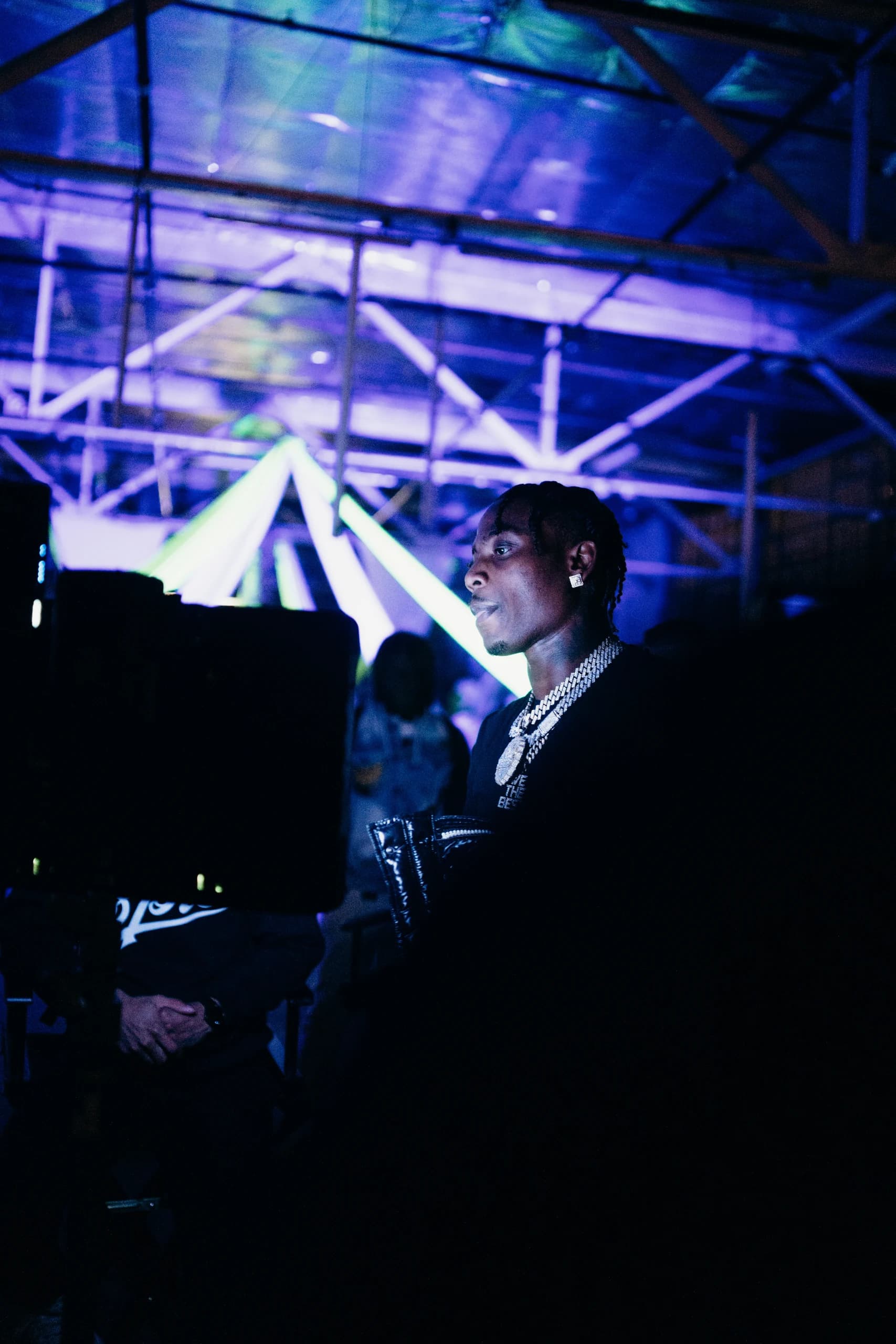 A performer beside a production monitor beneath blue and violet lights.