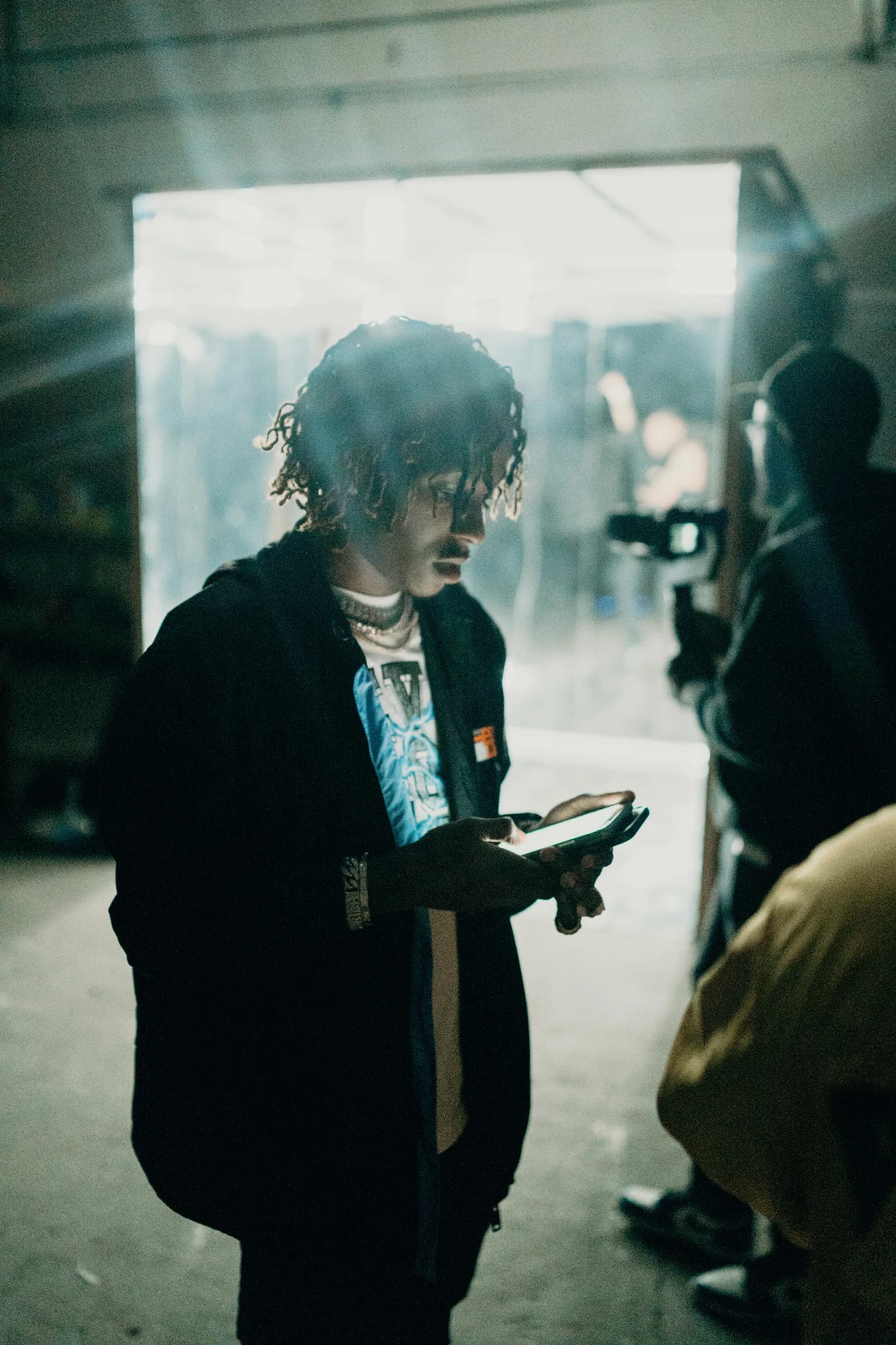 A performer checking his phone beside a camera operator and illuminated doorway.